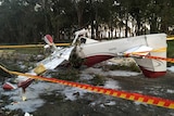The wreckage of a light plane sits on its roof on a dirt road in front of trees, surrounded by police tape.