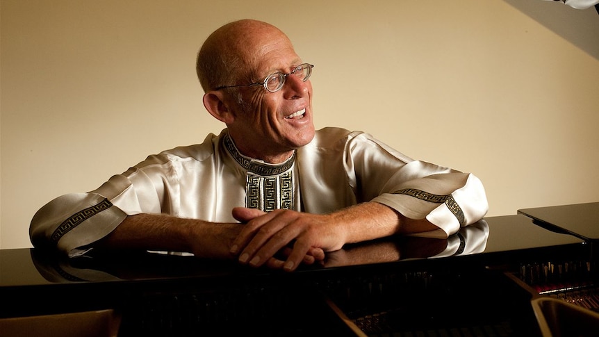 A older man wearing a white tunic and glasses sitting at a piano.