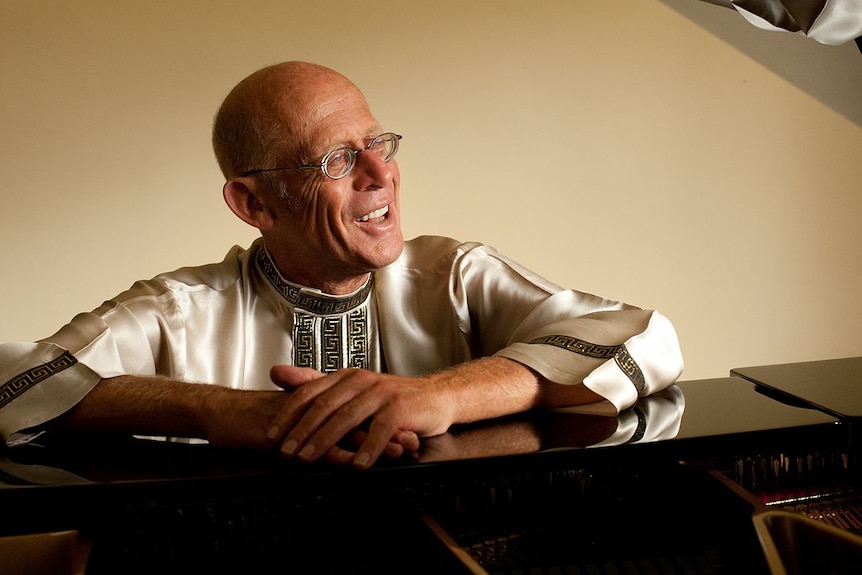 A older man wearing a white tunic and glasses sitting at a piano.