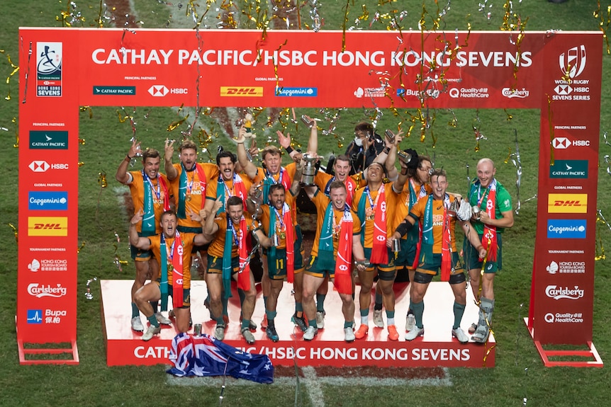 A group of players stand with streamers and confetti around them holding a trophy with a red advertising board around them