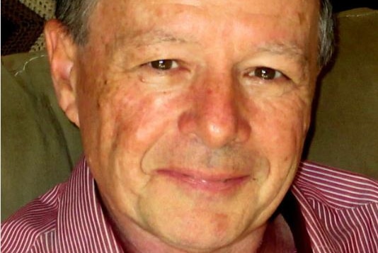 A close up photo of Tony wearing a red and white striped collared shirt and smiling with a closed mouth