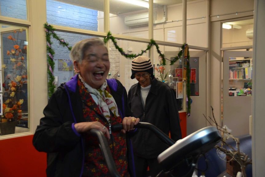 Two migrants laughing as one works out on a cross trainer. Decorations for Christmas in the background.