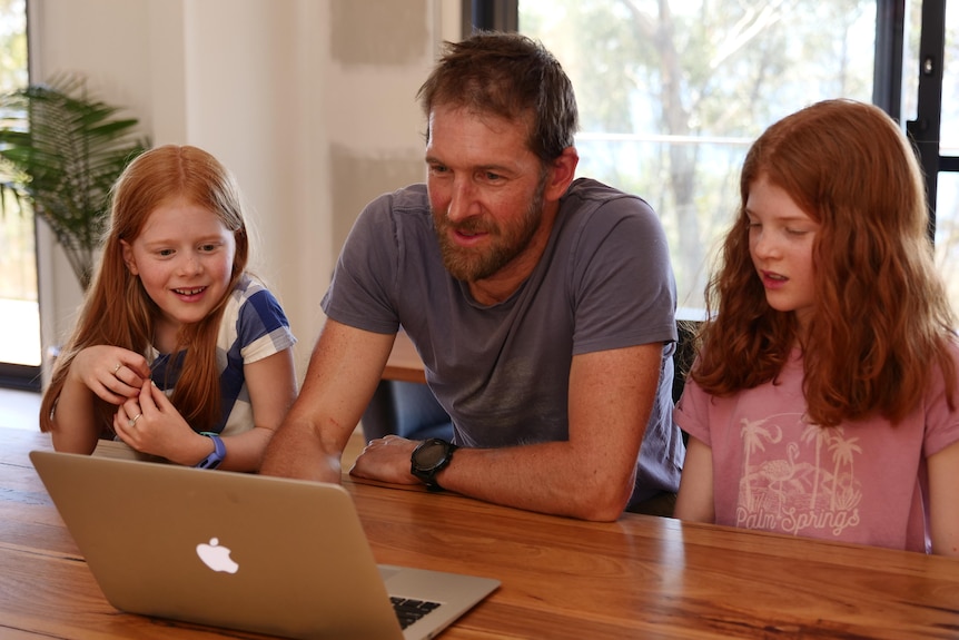 A man and his two young daughters look at a laptop screen.