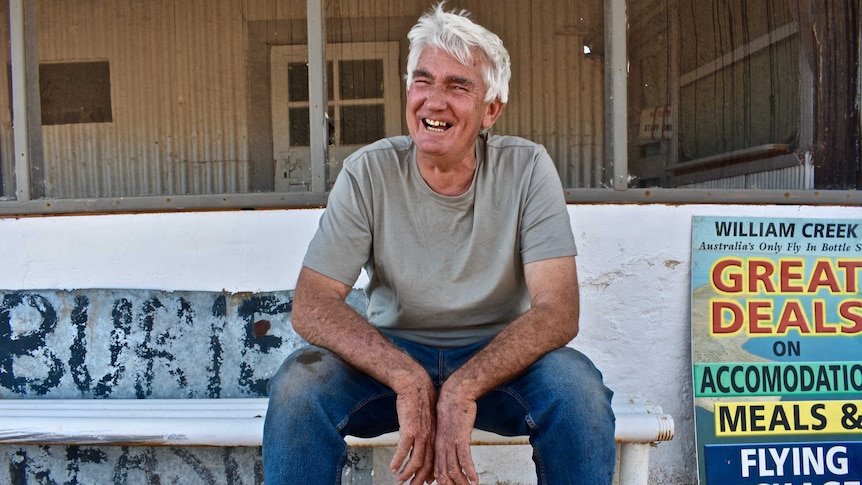 Man with white hair sits forward on an old white bench with a wide smile and looking straight down the camera