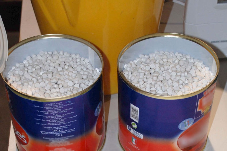 Two large tins labelled with tomatoes are filled with hundreds of tiny white pills.