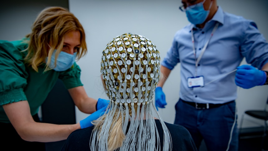 Fran wearing a cap with 250 little electrodes and wires running down her back, two medical professionals lean over her.