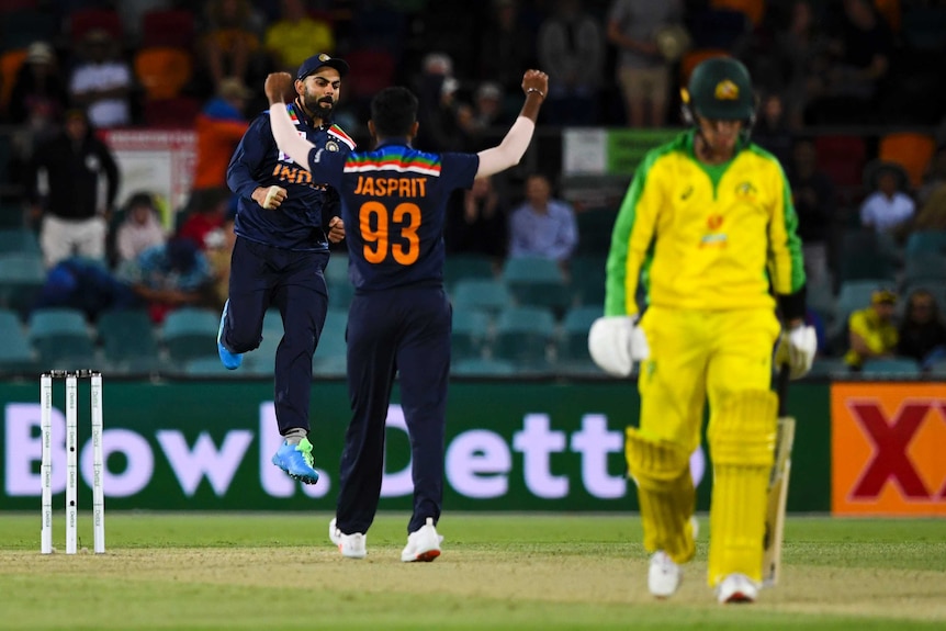 Two Indian male cricketers celebrate in the background as an Australia batter walks off the field.