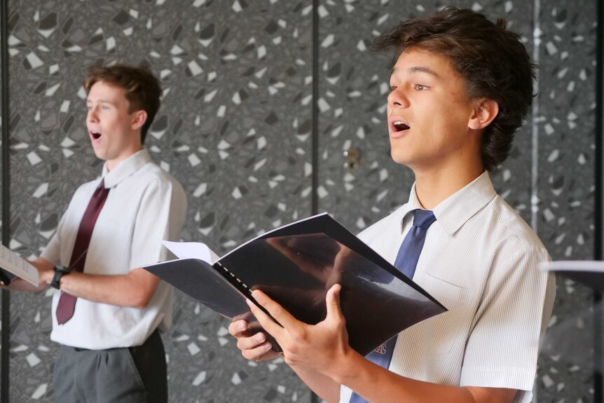 Two high school boys wearing white shirts and ties stand indoors singing.