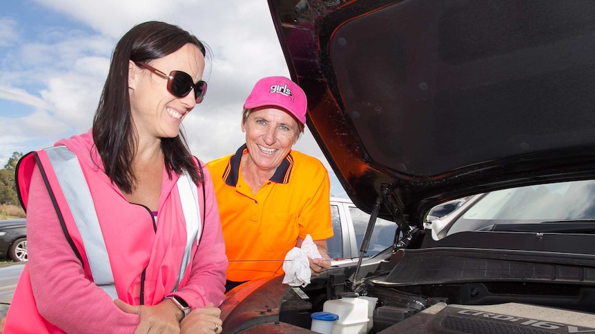 A woman in a pink high visibility vest and a woman in a bright orange shirt checking the oil on a car