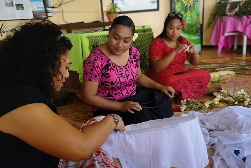 Three women sit on the ground and make nets and necklaces.