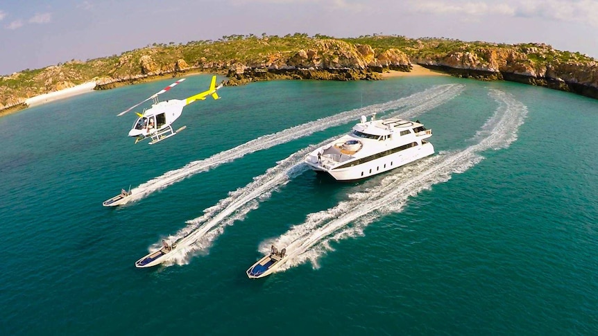 Three small boats move through emerald-green water, ahead of a larger cruise boat, while a helicopter flies overhead