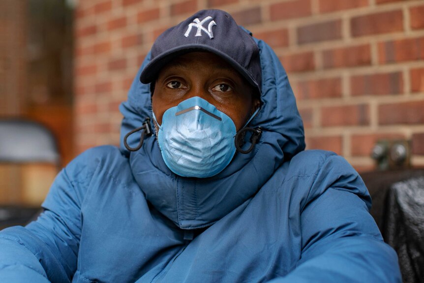A man in a cap, puffer jacket and face mask sitting on the side walk