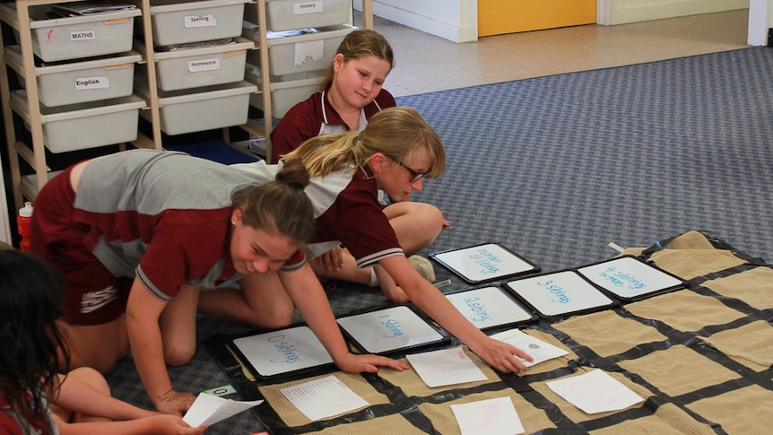 School children work with a large mat on the floor of their classroom.