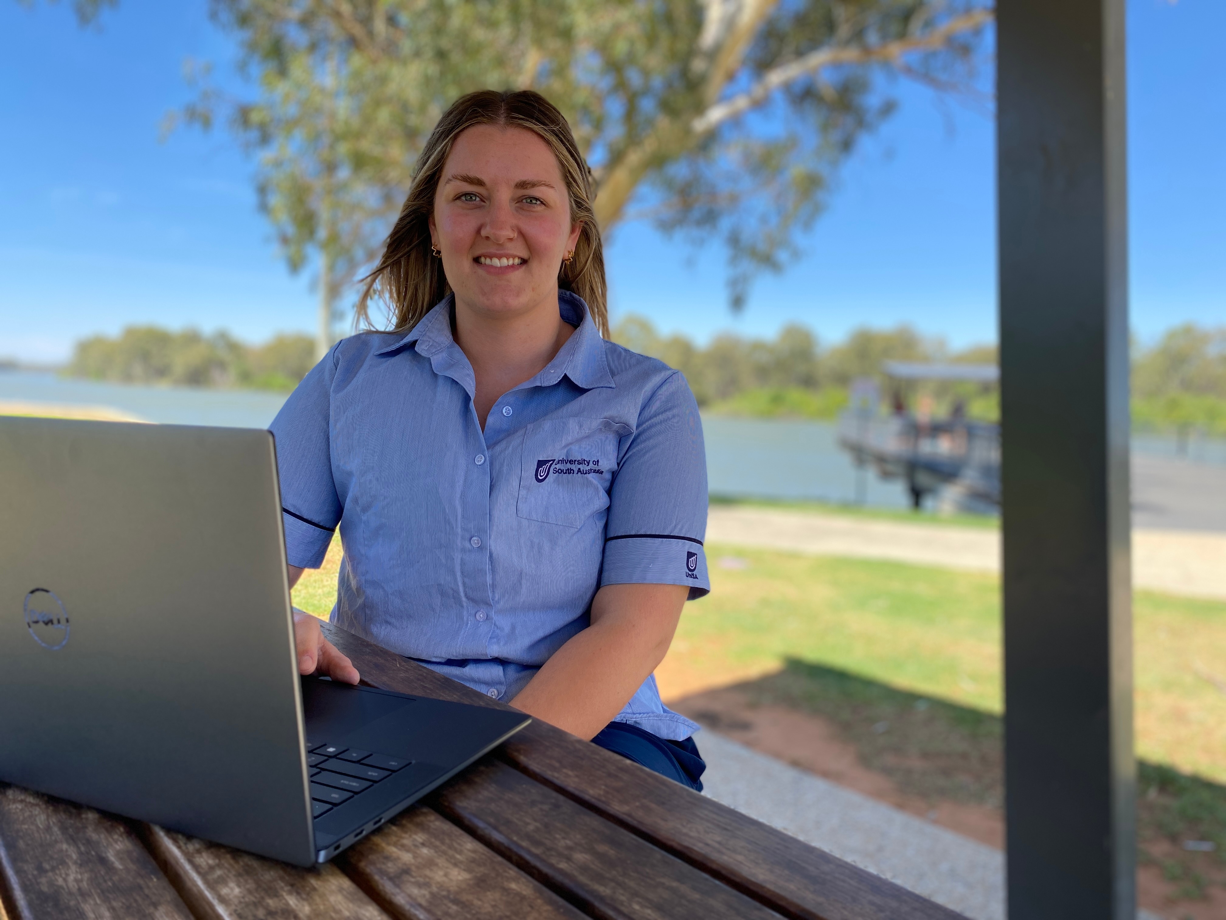 A woman with short blond hair smiles, she wears a blue university of south australia shirt, with a river in background