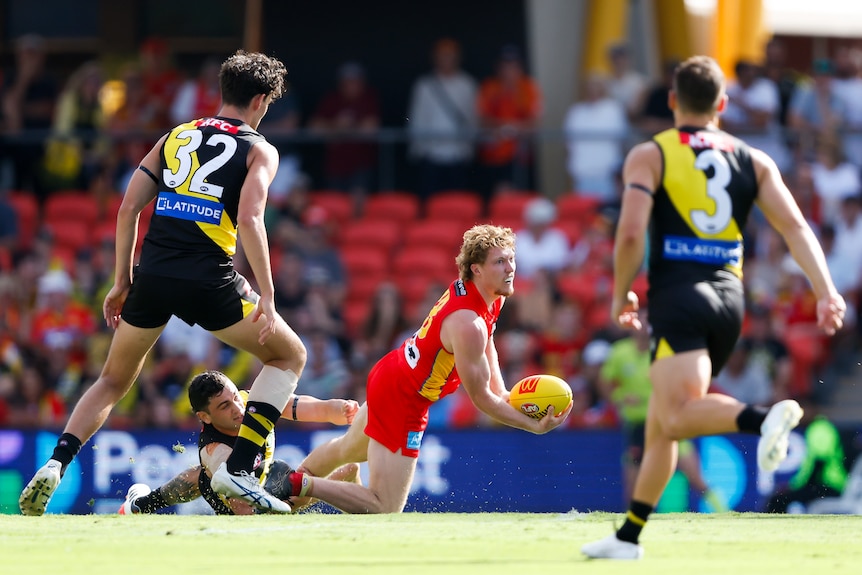 Gold Coast AFL player Matt Rowell handballs on his knees in the middle of the ground as Richmond players watch.