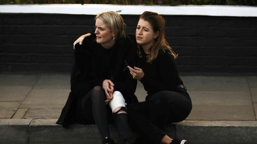 A woman with a bandage on her leg is consoled while sitting on the side of the street.