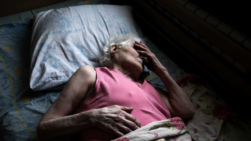 A woman wearing a pink singlet lies in bed holding her hand to her face.