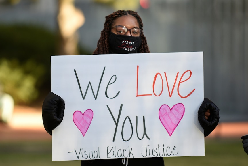 A young black woman wears face mask, hat and gloves and holds sign on cold day.