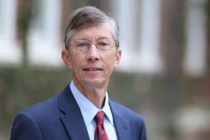 A man in a dark blue suit and red tie, and glasses, smiles slightly before a blurred background.