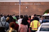 A group of people in quad listen to people speaking next to brick wall saying "Richnick Elementary School"