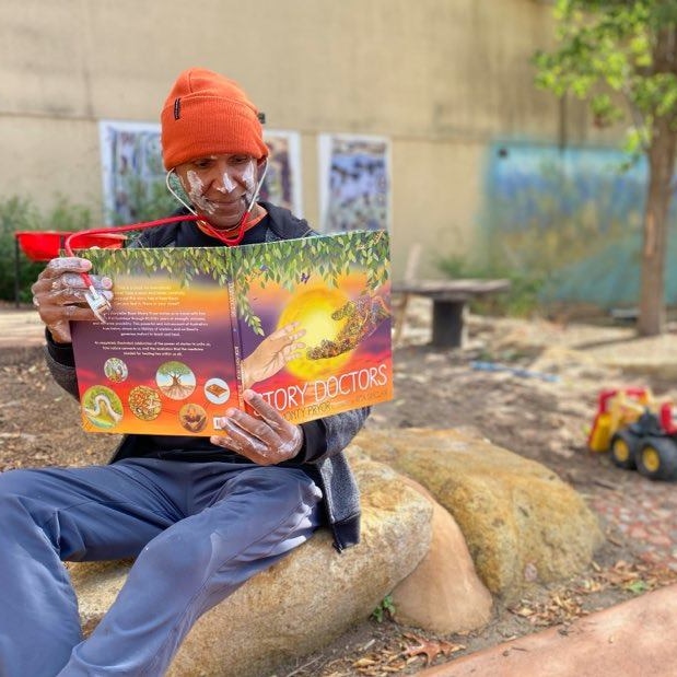 Indigenous man with white paint on wearing an orange beanie and a stethoscope while reading a book on a rock