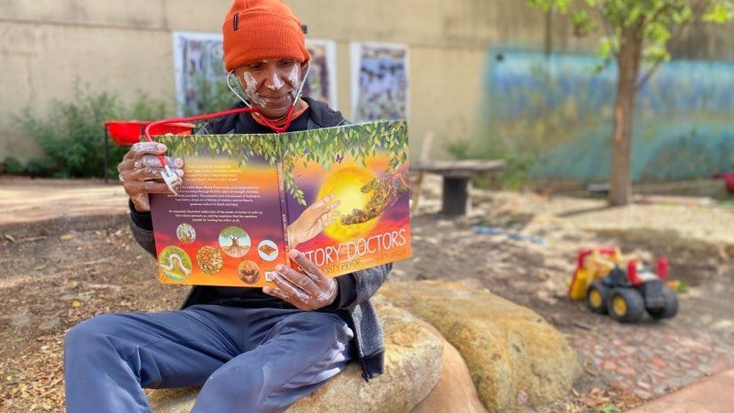 Indigenous man with white paint on wearing an orange beanie and a stethoscope while reading a book on a rock