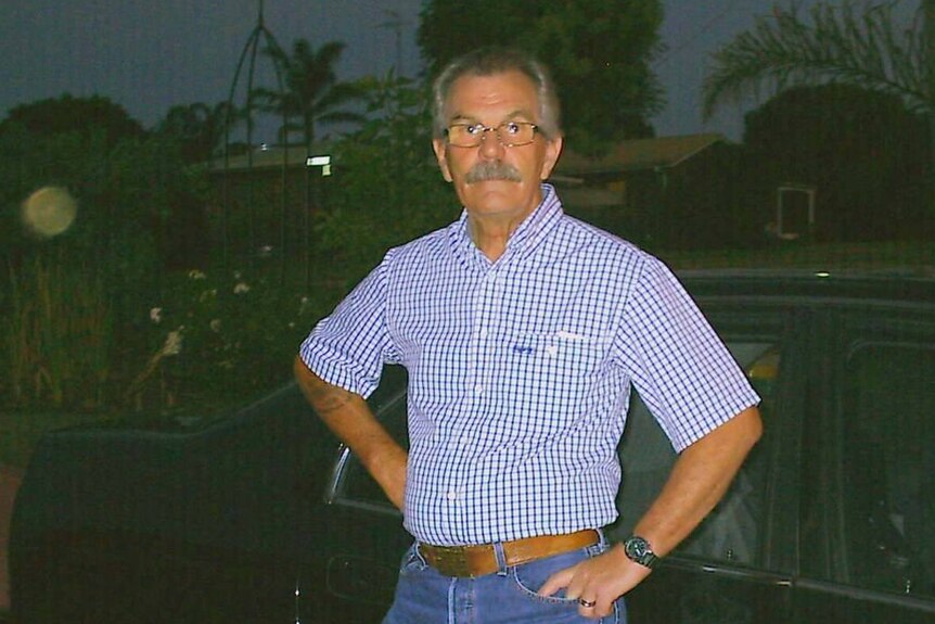 A mature gentleman in jeans, button up shirt and white shoes stands in front of a car.
