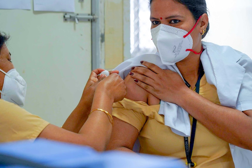 A woman in a face mask and bindi holding her shirt down while she is injected by a nurse