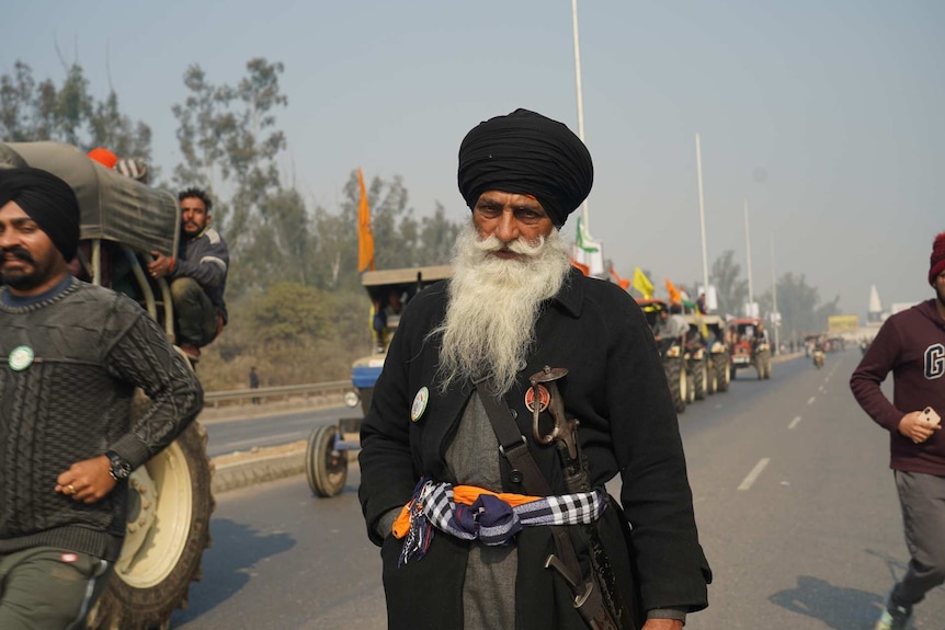 a man dressed in black with a sword looks at the camera as men on tractors drive beside him on the road