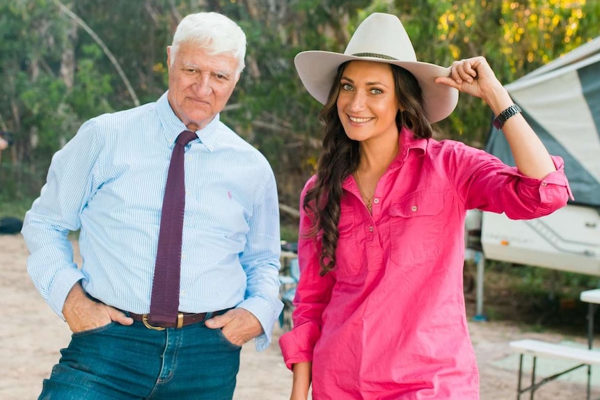 A woman in a bright pink shirt tips her cowboy hat and smiles at the camera alongside an old man with white hair.