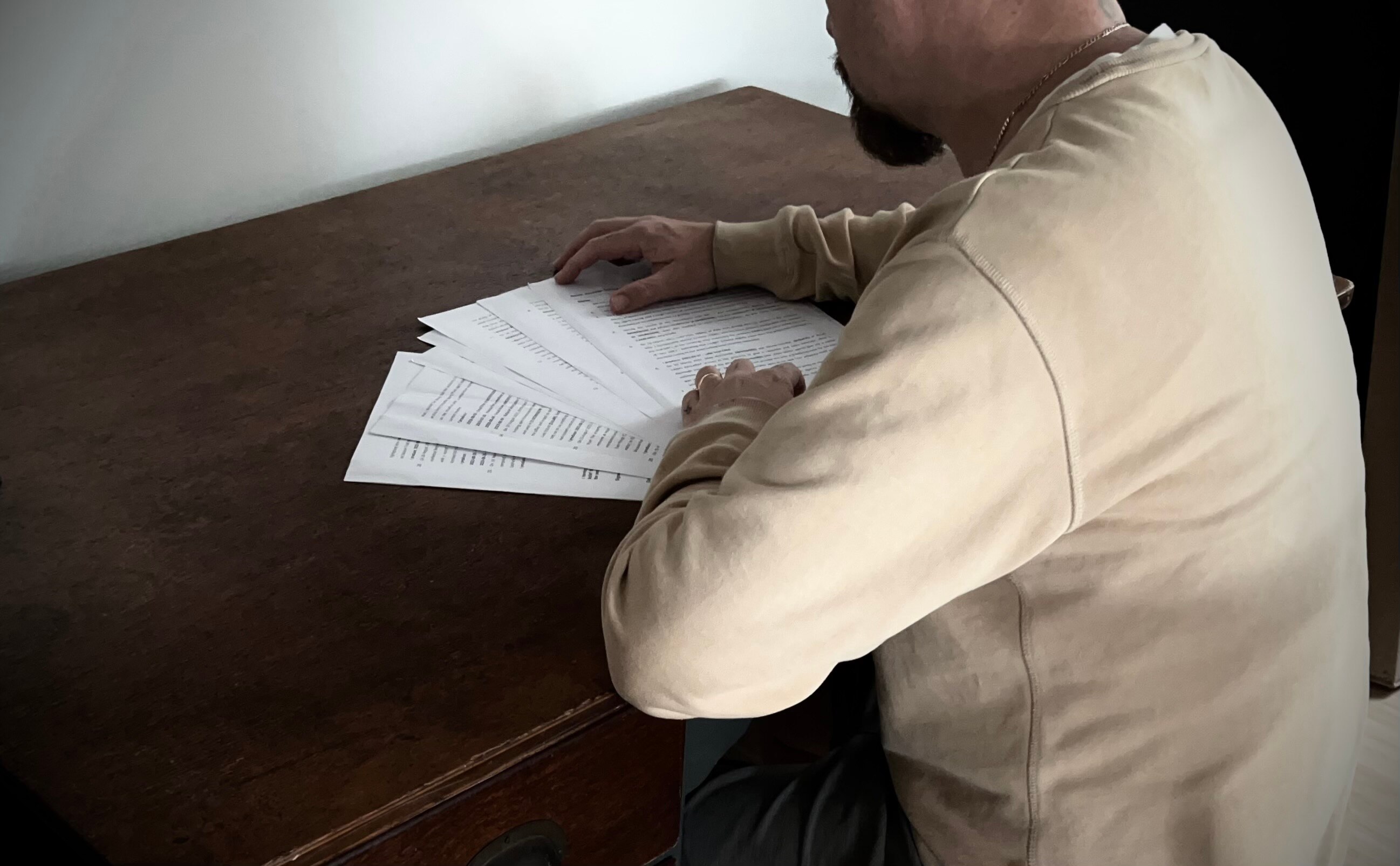 A man with a short, dark beard, wearing a beige top, sitting at a table with some papers.