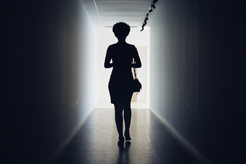 Rear view of a silhouetted young businesswoman walking down a dark office corridor to a lit doorway.