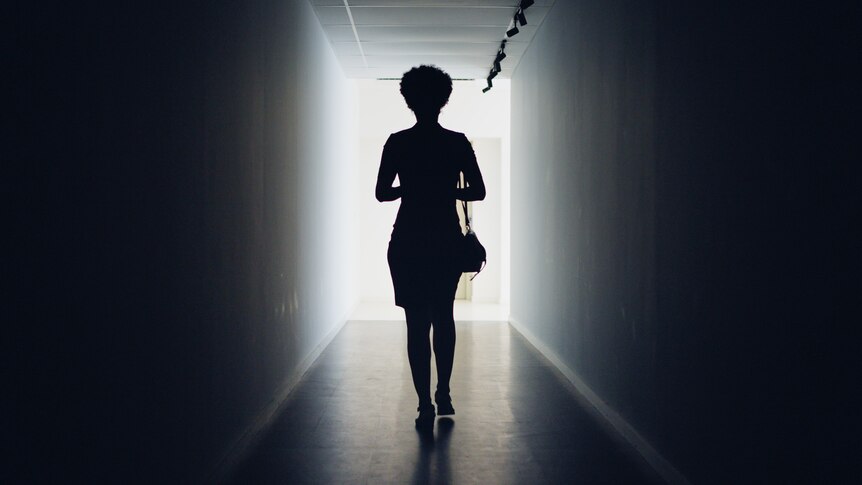 Rear view of a silhouetted young businesswoman walking down a dark office corridor to a lit doorway.