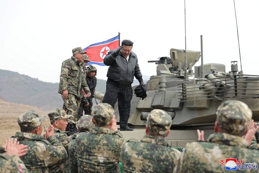 Kim Jong Un stands on top of a tank looking down as soldiers stand below.
