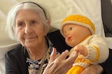 An elderly woman with white hair holds a baby doll wearing a yellow beanie.