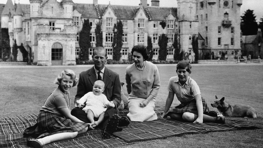 A black and white photo of Queen Elizabeth and Philip sitting on a picnic blanket with their kids and dog