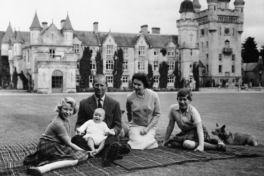 A black and white photo of Queen Elizabeth and Philip sitting on a picnic blanket with their kids and dog