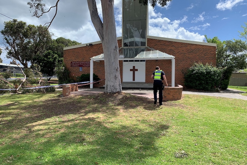 A policeman stands on grass outside a parish in Parkdale with a fallen branch on his left cordoned off by police tape.