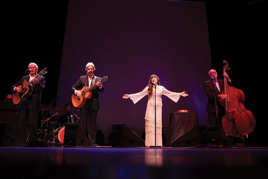 A woman in a white dress and three men in tuxedos on stage, playing the role of The Seekers.