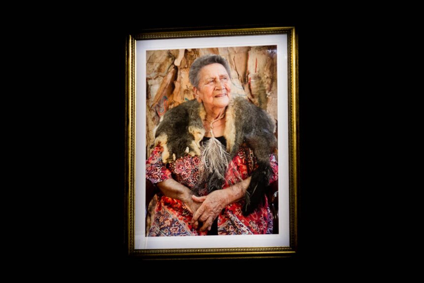 A gold-framed portrait of an older woman wearing a colourful dress and an animal skin around her neck hangs on a black wall.