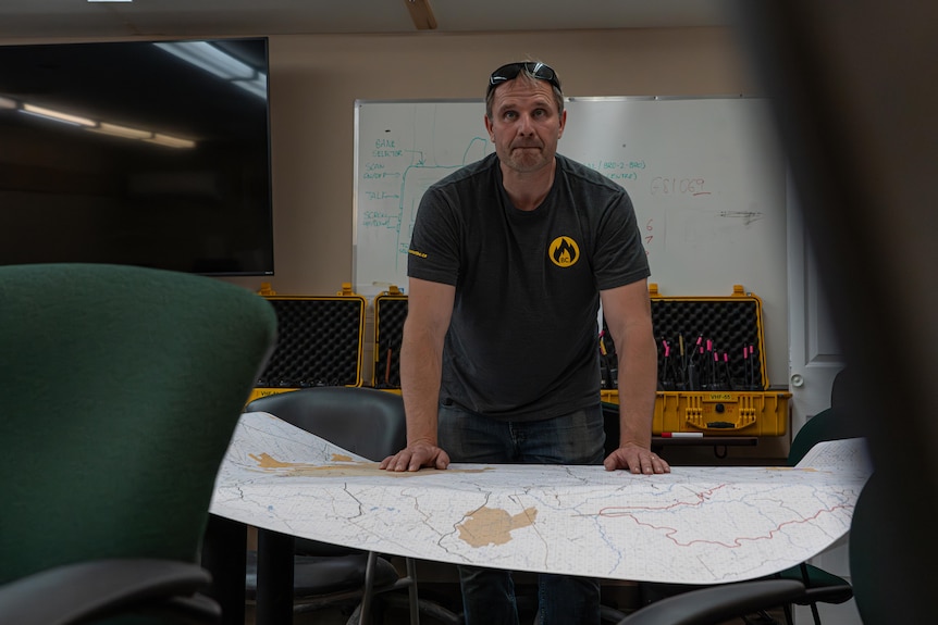 A man stands over a table with a large map on it. He has both his hands on the map, leaning on it, and looks towards the camera.