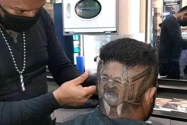 A man with a picture of a face shaved into his hair sits beside a hairdresser.