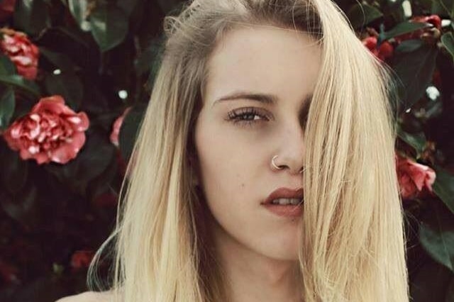 An androgynous model with long blond hair standing in front of a red camelia bush