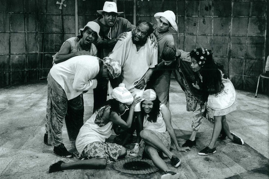 A black and white picture of nine actors in a huddle on stage during a performance of the Jimmy Chi play Corrugation Road.