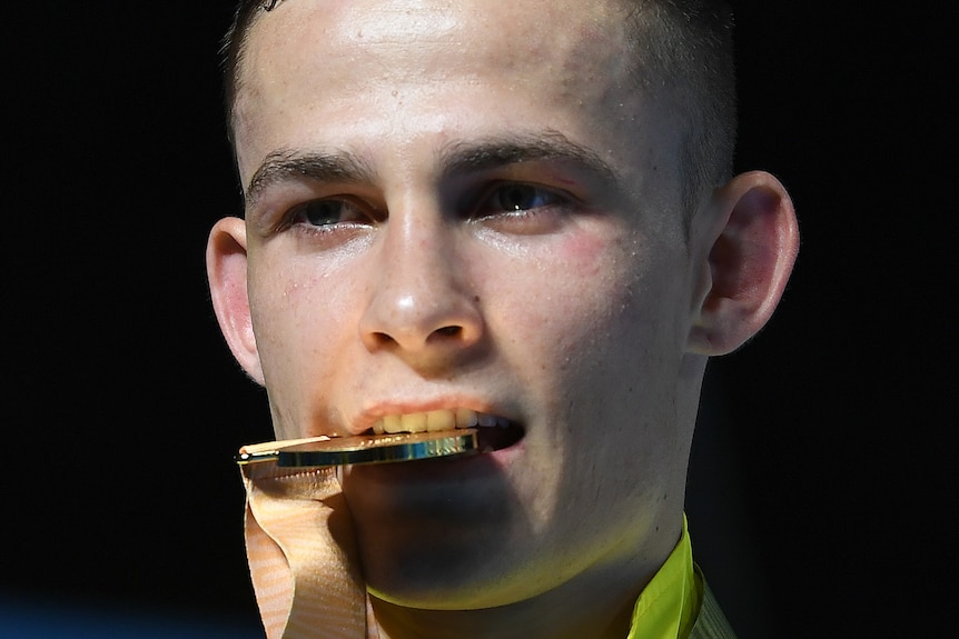 Young, brunette man holds gold medal in teeth