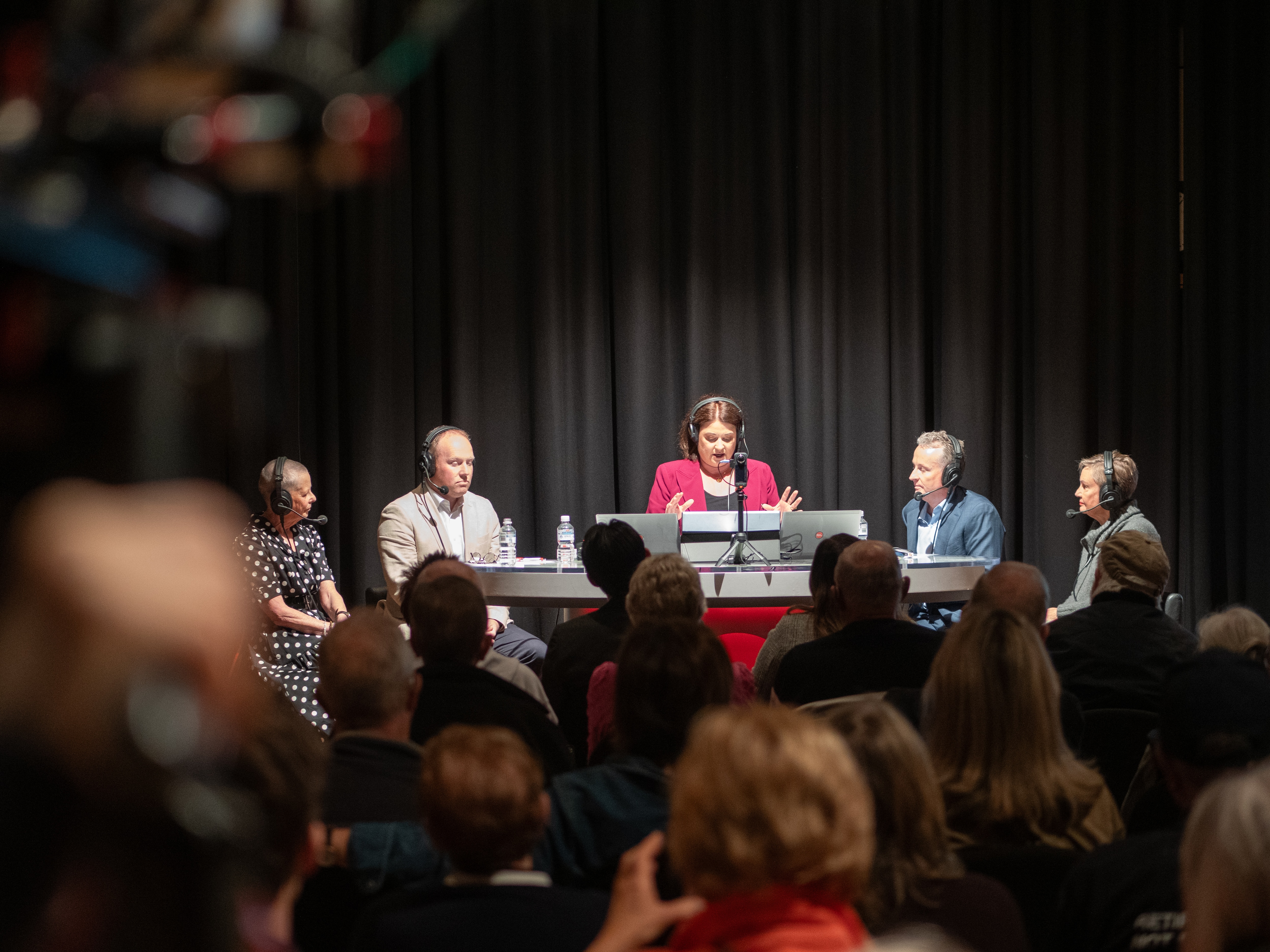 Radio panel of five people sat at desk in front of studio audience