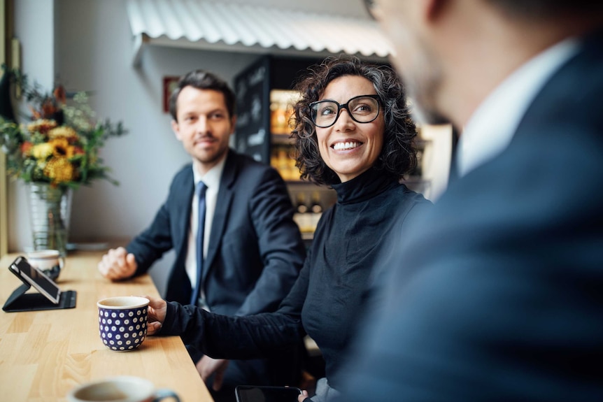 Two men and a woman sit at a table in a work environment conversing over coffee.