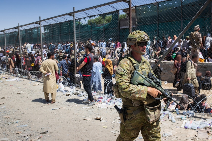 A soldier passes wire fencing behind which crowds of people stand.