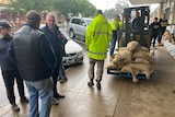 People standing on a footpath, as a machine carries sandbags.