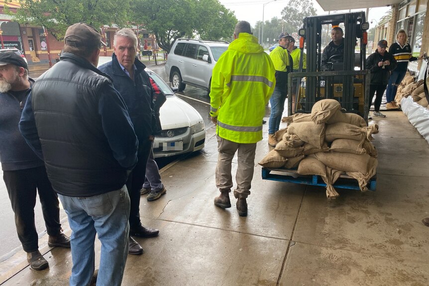 People standing on a footpath, as a machine carries sandbags.